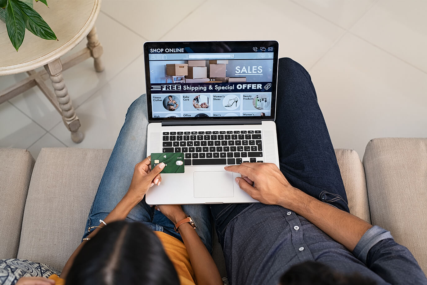 Two people sitting on a couch shopping online, one holding a credit card while browsing a laptop displaying sales, free shipping, and special offer promotions.